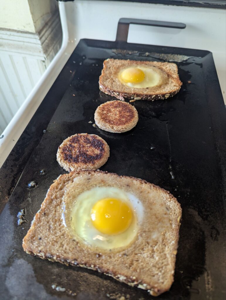 A griddle on top of a gas range. There are two pieces of bread on top. Each has a small circle taken out of it. The circles are on the griddle next to the bread. In the hole where the circles were are eggs cooking