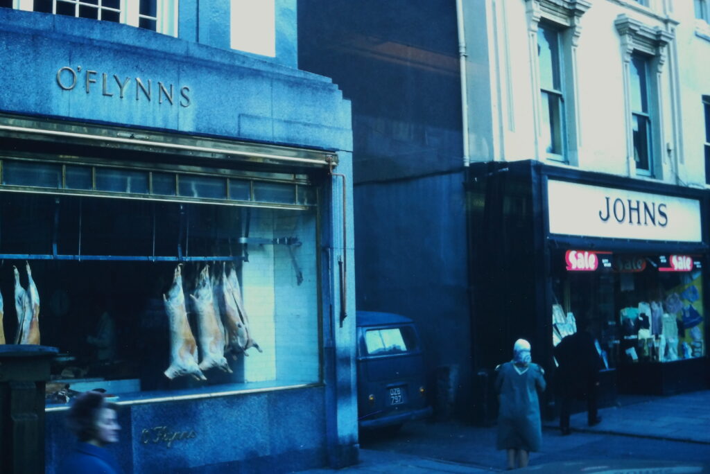 Butchered animal carcasses hang in the window of a place called "O'Flynn's". Next door is a clothing store called "Johns"