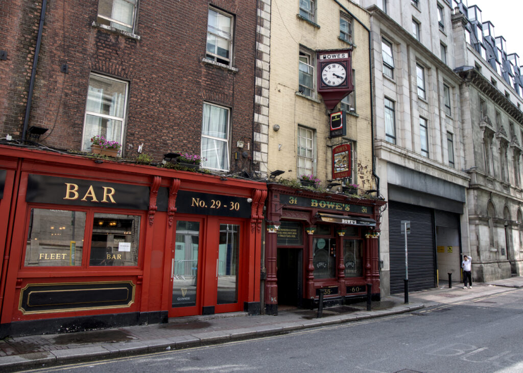 A row of buildings. The first one is a bar painted bright red. Next is Bowe's. There is a man with his hand on his head in front of a roll up door beyond Bowe's
