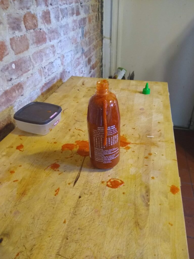 Bottle of Sriracha on the cutting board. The seal is nowhere to be seen. Sriracha has spilled