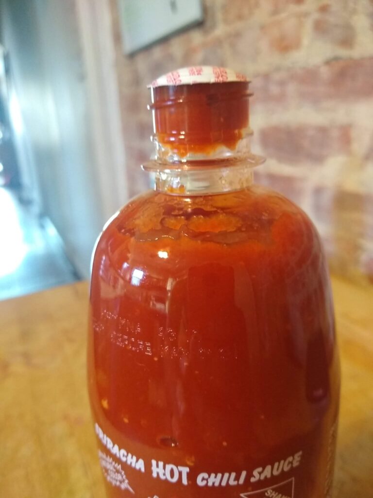 A bottle of Sriracha. The cap is off but the seal is still on. It is bulging ominously
